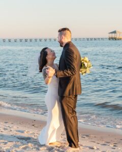 Bride and groom smiling barefoot on sandy beach, holding hands by ocean and pier at Gulf South wedding.