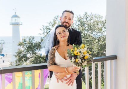 Bride and groom smile on a balcony with bouquet, lighthouse, and trees in sunny Louisiana wedding setting.