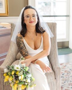 Bride with glasses and floral arm tattoo smiles in striped chair, holding yellow-white bouquet at Louisiana wedding.