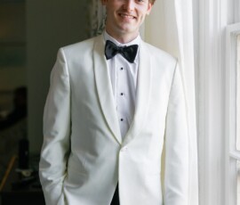 Young man in a white tuxedo jacket and black bow tie stands by a window, smiling—classic wedding style.