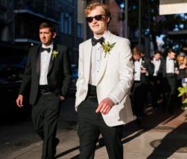 Man in white tuxedo and sunglasses walks confidently with others in black tuxedos, exuding New Orleans wedding style.