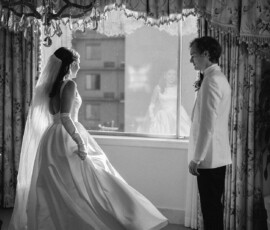 Bride in white gown and veil stands by a window, facing groom; her reflection appears in this elegant black and white wedding photo.