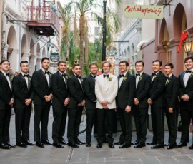 Twelve men, eleven in black tuxedos and one in white, smile together outdoors, likely at a Louisiana wedding.