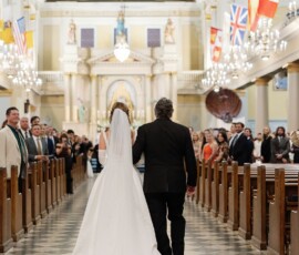 Bride in white gown walks down church aisle with older man, surrounded by guests, chandeliers, and international flags.