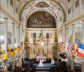 A New Orleans wedding ceremony in an ornate church with vaulted ceilings, chandeliers, murals, and flags; couple at the altar.