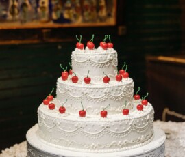 Three-tiered white cake with icing swirls and cherries on a silver stand, set on a white tablecloth at a Gulf South wedding.