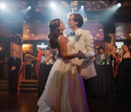 Bride and groom share their first dance in a warmly lit Gulf South venue as guests watch from the floor and balcony.