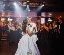 Bride and groom share a romantic first dance in a dimly lit venue, watched by elegantly dressed guests on floor and balcony.