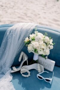 Bridal bouquet, pearl heels, BRIDE clutch, sunglasses, and pearl veil on blue couch with sand in background.