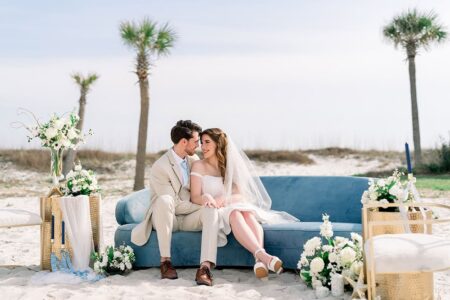 Bride and groom smile at each other on a blue couch at a beach wedding, surrounded by white flowers and palm trees.