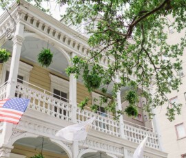 Charming two-story pale yellow house with white trim, railings, hanging ferns, flags, and leafy tree shade—ideal Louisiana wedding setting.