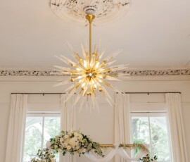 Gold starburst chandelier over floral wedding arch in a bright room with tall windows and white curtains.