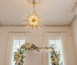 Gold starburst chandelier above floral wedding arch with white flowers, greenery, and tall curtained windows in Louisiana.