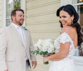 Bride in white off-shoulder dress smiles at groom in beige suit outside cream building with large windows.