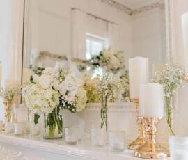 White and gold candles, white flowers, and glass vases decorate a mantel in front of a mirror—classic Louisiana wedding decor.