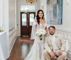 Bride in lacy white gown with bouquet stands by seated groom in beige suit, bright hallway, wood floors, wedding inspiration.