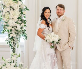 Bride in lace gown and groom in beige suit smile under a white floral arch, holding a bouquet—Gulf South wedding inspiration.