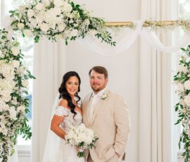 Bride in off-shoulder gown and groom in beige suit smile under a floral arch with white flowers and greenery.