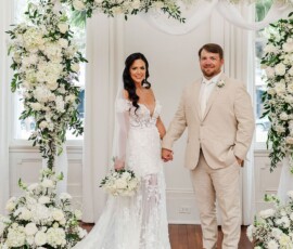 Bride in white lace gown and groom in beige suit hold hands under a floral arch at a bright wedding ceremony.