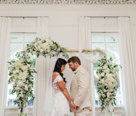 Bride and groom hold hands under a white floral arch in a bright New Orleans wedding room with large windows and white curtains.