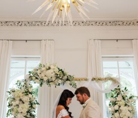 Bride and groom hold hands under a floral arch in a bright, elegant room with white curtains and a gold chandelier.