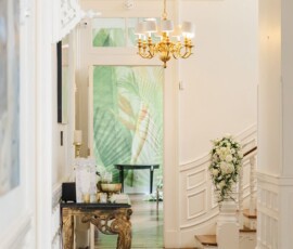 White-walled hallway with gold chandelier, floral staircase, and leafy mural—ideal Louisiana wedding venue.