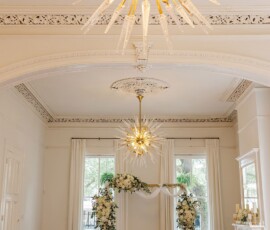 Bright indoor wedding venue with white chairs, floral decor, gold chandeliers, and airy windows for a timeless Gulf South ceremony.