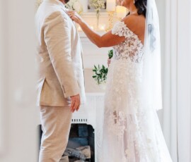Bride in floral lace gown adjusts groom’s lapel as they smile at each other by a flower-adorned fireplace in a bright room.