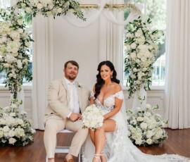 Bride in white dress and groom in beige suit sit indoors with white flowers, large windows, and a classic Louisiana wedding feel.