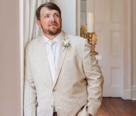 Man in a beige suit and white tie looks out a sunlit window, candles in hallway behind him, awaiting the wedding ceremony.