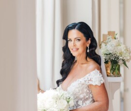 Bride with long dark hair in a white lace gown holds a white bouquet, smiling by a window with floral Gulf South wedding vibes.