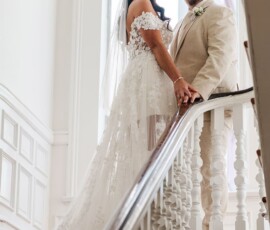 Bride in white lace gown and groom in beige suit hold hands, smiling on a sunlit staircase at a Louisiana wedding.