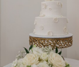 Elegant two-tier white wedding cake with florals on a gold stand and white rose bouquet, perfect for Louisiana weddings.
