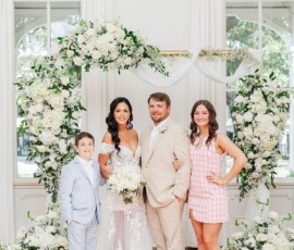 Bride, groom, and two kids smile before a heart-shaped floral display at their joyful New Orleans wedding ceremony.