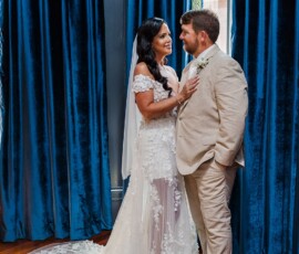 Bride in white lace gown and groom in beige suit share a smile by blue-curtained window; bouquet on floor inspires wedding style.
