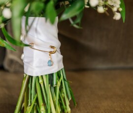 Wedding bouquet with green stems, white ribbon, gold pin and blue charm, topped with green leaves and white flowers.