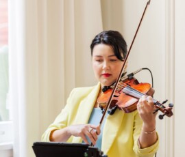 Woman in yellow blazer plays violin indoors by window, music stand and electronic gear evoke elegant Louisiana wedding ambiance.