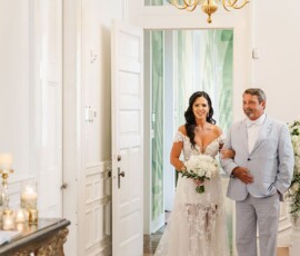 Bride in white gown with bouquet walks arm-in-arm with older man into bright, flower-filled New Orleans wedding ceremony.