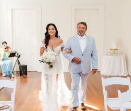 Bride in white and older man walk down the aisle at a Louisiana wedding; violinist plays, with white chairs and cake table nearby.