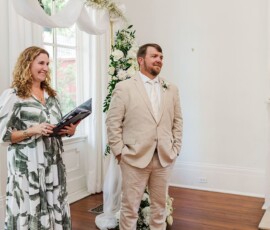 Man in beige suit smiles beside woman with black folder by floral arch and white fireplace at indoor wedding ceremony.