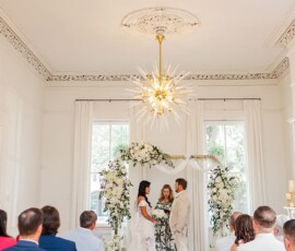 Bride and groom exchange vows under a floral arch at a bright, elegant Louisiana indoor wedding with guests seated nearby.