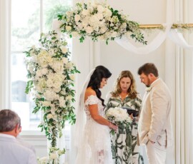 Bride and groom hold hands under a floral arch as an officiant reads, with guests watching a romantic Gulf South wedding.