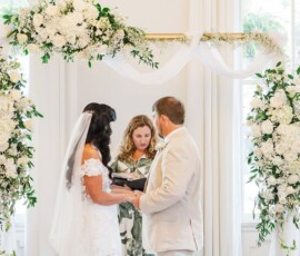 Bride and groom hold hands under a floral arch as an officiant reads during their indoor Gulf South wedding ceremony.