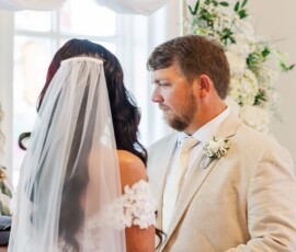 Bride in veil and lace dress holds hands with groom in beige suit at indoor Louisiana wedding, white flower decor behind them.