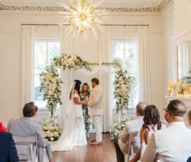 A couple exchanges vows under a floral arch in a bright room as guests watch a Gulf South-inspired wedding ceremony.