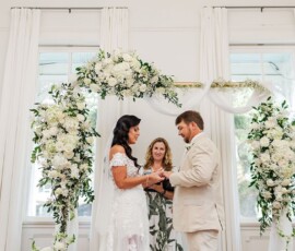Couple exchanges rings indoors under a white floral arch, officiant smiling—classic Louisiana wedding elegance.