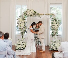 Bride and groom exchange vows under a floral arch indoors, with guests watching in an elegantly decorated, bright wedding room.