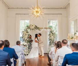 Couple exchanges vows under a floral arch at a Louisiana wedding, with guests seated in a bright, elegant room.
