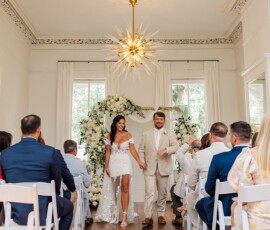 Bride in white dress and groom in beige suit walk aisle, smiling, as guests watch in a bright, floral New Orleans wedding.