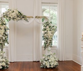 Elegant white floral wedding arch with greenery in a bright room with tall windows, white curtains, and wooden floors.
